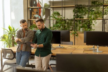 İki profesyonel, çağdaş bir ofis alanında dijital tablet tutarken, kırsal tuğla ve sıcak doğal ışık içeren odaklı bir sohbete girişiyor.