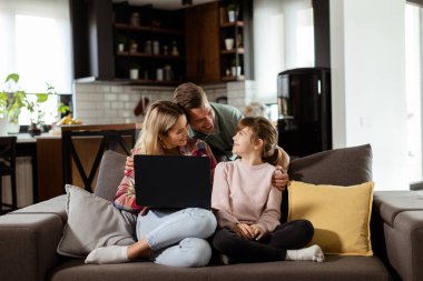 Üç kişilik neşeli bir aile oturma odasındaki kanepede birlikte vakit geçirir, rahat evlerinde dizüstü bilgisayarın etrafında bir an paylaşırlar.