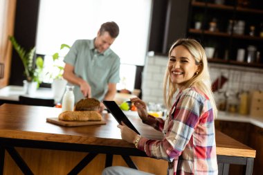 Kadın mutfak tezgahında elinde domates tutan bir adam ona gülümserken bir tabletten okuyor.