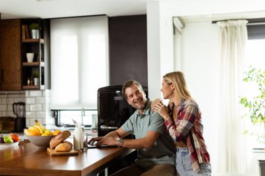 Neşeli bir çift güneşli mutfaklarında keyifli bir anın tadını çıkarıyor. Sağlıklı bir kahvaltıyla çevrili bir laptopla çalışıyorlar.