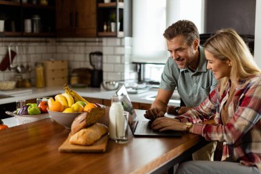 Neşeli bir çift güneşli mutfaklarında keyifli bir anın tadını çıkarıyor. Sağlıklı bir kahvaltıyla çevrili bir laptopla çalışıyorlar.