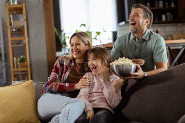Üç kişilik bir aile rahat bir şekilde kanepeye kurulmuş, heyecanlı bir film gecesinde bir kase patlamış mısırı paylaşırken yüzlerindeki heyecanı ve dikkati yansıtıyor.