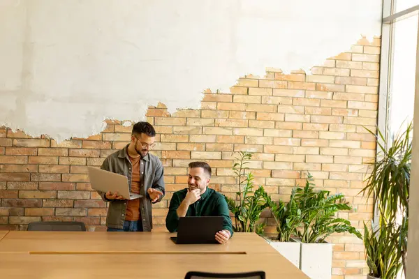 stock image Two smiling professionals engage in a collaborative work session at a wooden table, their camaraderie evident in a contemporary office setting with an exposed brick wall backdrop