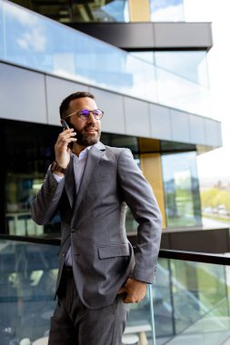 Kollarını kavuşturmuş uygun bir adam, parlak bir binanın önünde gururla duruyor, hırsını yansıtıyor..