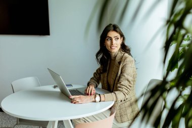 Süslü ceketli, şık bir iş kadını laptopuyla beyaz bir masada minimalist dekorlarla çevrili..