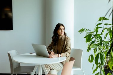 Süslü ceketli, şık bir iş kadını laptopuyla beyaz bir masada minimalist dekorlarla çevrili..