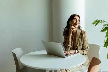 Bir kadın, güneş ışığında beyaz bir masada oturuyor. Düşüncelere dalmış, yanında bir laptop ve yeşil bitkiyle..