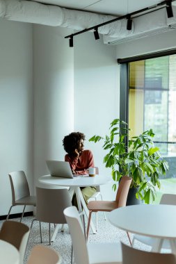 Afro-Amerikalı bir kadın şık bir kafede oturuyor, önünde dizüstü bilgisayarıyla içkisini yudumluyor, gün ışığında güneşleniyor.