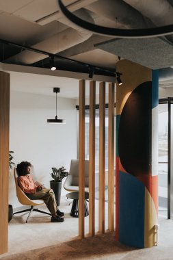 Afro-Amerikalı bir kadın, canlı bir sörf tahtası ve yemyeşil bir saksı bitkisinin yanında akıllı telefonlarına dalmış, bir sandalyede dinleniyor..