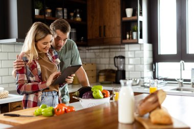 Neşeli bir çift, taze malzemelerin arasında dijital tablete sarılmış, iyi aydınlatılmış bir mutfakta duruyor.