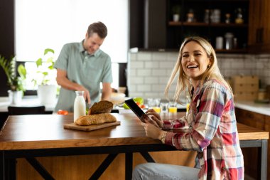 Kadın mutfak tezgahında elinde domates tutan bir adam ona gülümserken bir tabletten okuyor.