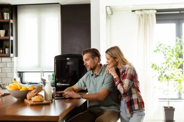 Neşeli bir çift güneşli mutfaklarında keyifli bir anın tadını çıkarıyor. Sağlıklı bir kahvaltıyla çevrili bir laptopla çalışıyorlar.