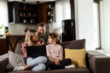 Üç kişilik neşeli bir aile oturma odasındaki kanepede birlikte vakit geçirir, rahat evlerinde dizüstü bilgisayarın etrafında bir an paylaşırlar.