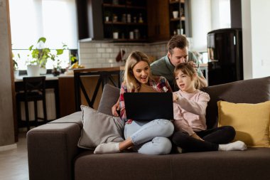 Üç kişilik neşeli bir aile oturma odasındaki kanepede birlikte vakit geçirir, rahat evlerinde dizüstü bilgisayarın etrafında bir an paylaşırlar.
