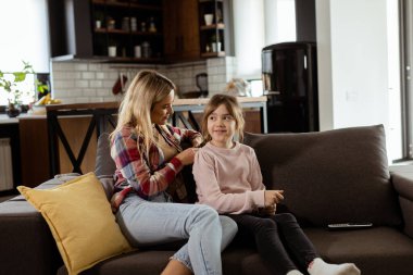 Nazikçe dokunmak, sessiz bir sabahı zenginleştirir. Bir anne, kızının saçlarını karmaşık bir şekilde oturma odasındaki kanepede örer.