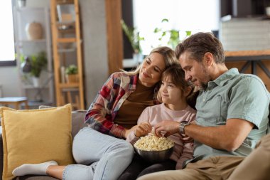 Üç kişilik bir aile rahat bir şekilde kanepeye kurulmuş, heyecanlı bir film gecesinde bir kase patlamış mısırı paylaşırken yüzlerindeki heyecanı ve dikkati yansıtıyor.