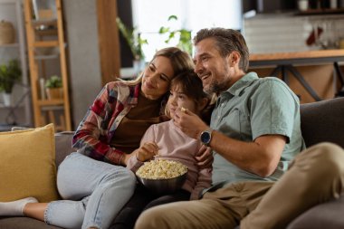 Üç kişilik bir aile rahat bir şekilde kanepeye kurulmuş, heyecanlı bir film gecesinde bir kase patlamış mısırı paylaşırken yüzlerindeki heyecanı ve dikkati yansıtıyor.