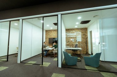 İki profesyonel, yeşil ve çağdaş tasarım unsurlarıyla çevrili bir ofis salonunda rahatça otururken sohbet ediyorlar.