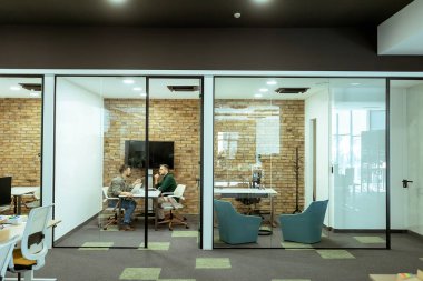 İki profesyonel, yeşil ve çağdaş tasarım unsurlarıyla çevrili bir ofis salonunda rahatça otururken sohbet ediyorlar.