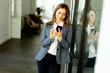 Ofis koridorunda neşeli, profesyonel bir kadın duruyor. İçerik yansıması bir akıllı telefonu tutarken görülüyor..