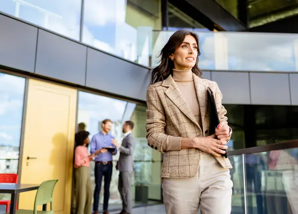 質素なプロの女性がガラス張りのオフィススペースを歩き回り 同僚が背景で会話する — ストック写真
