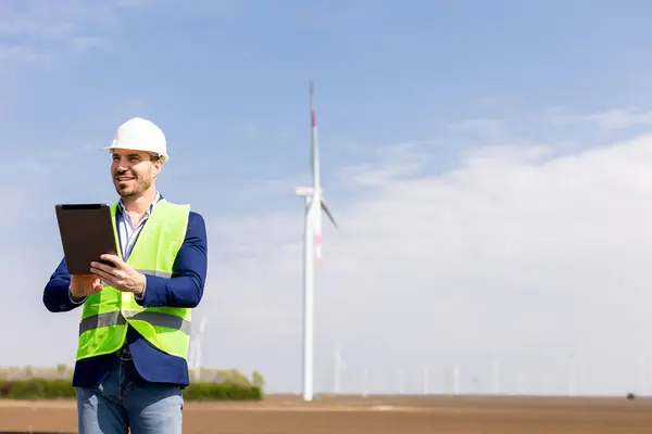 A cheerful engineer in safety gear reviews data on a tablet before towering windmills under a clear sky.