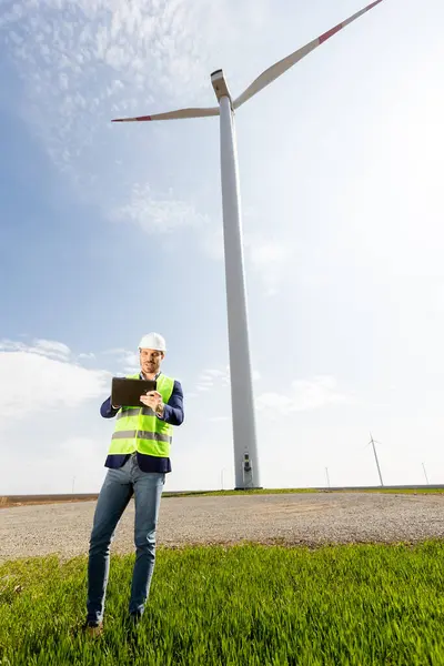 A safety-helmeted engineer with clipboard scrutinizes the workings of a majestic wind turbine amidst lush greenery under a clear sky.