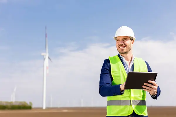 A cheerful engineer in safety gear reviews data on a tablet before towering windmills under a clear sky.