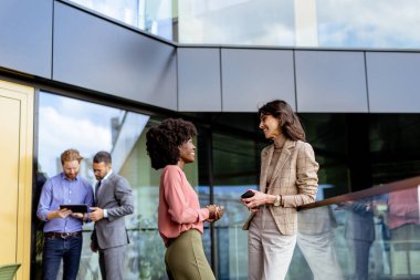 İki kadın çok ırklı meslektaşı ofis dışında arkadaşça sohbet ederken, diğerleri arka planda çalışıyor.