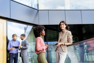 İki profesyonel kadın hareketli bir şekilde sohbet ederken, erkek meslektaşlar arka planda tartışıyorlar..