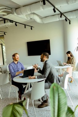 Bir kadın arka planda bağımsız olarak çalışırken, iki erkek bir masada canlı bir konuşma yapar.