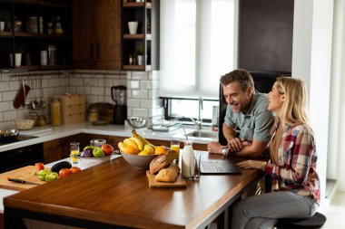 Neşeli bir çift güneşli mutfaklarında keyifli bir anın tadını çıkarıyor. Sağlıklı bir kahvaltıyla çevrili bir laptopla çalışıyorlar.