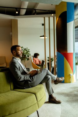 Odaklanmış iş adamı, canlı, çağdaş bir çalışma alanında rahat yeşil bir koltukta otururken dizüstü bilgisayarı üzerinde çalışır..