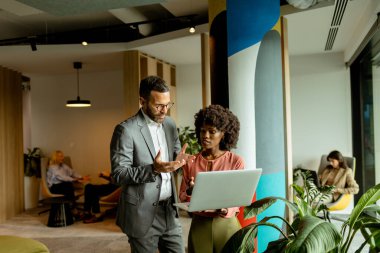 Erkek ve kadın, arka planda meslektaşlarıyla canlı bir ofis ortamında dizüstü bilgisayarda iş tartışması yapıyorlar.