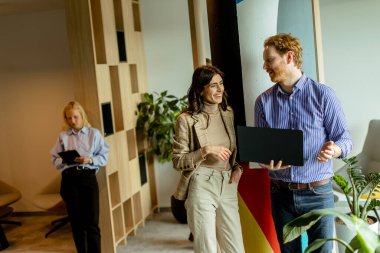 İki profesyonel, arka planda özenli bir meslektaşla fikir alışverişinde bulunarak bir dizüstü bilgisayarda işi tartışıyor.