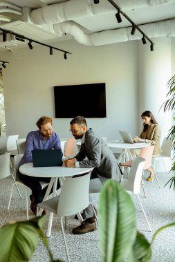 Bir kadın arka planda bağımsız olarak çalışırken, iki erkek bir masada canlı bir konuşma yapar.