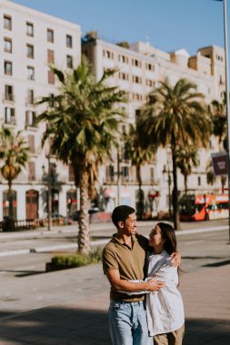 Genç çift Barselona 'da avuçlar tarafından çerçevelenmiş güneşli bir yolda yürürken el ele tutuşup gülümsüyor.