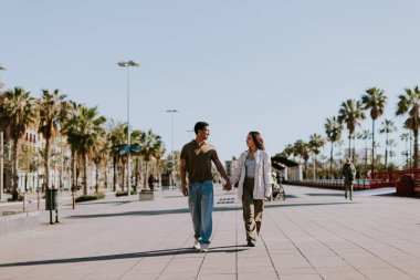 Genç çift Barselona 'da avuçlar tarafından çerçevelenmiş güneşli bir yolda yürürken el ele tutuşup gülümsüyor.