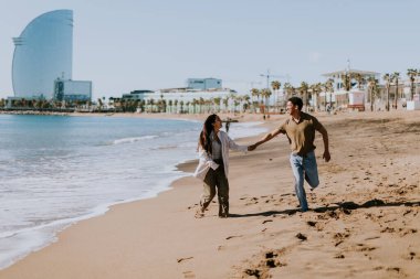 Gülümseyen bir çift kumlu bir sahilde kaygısızca koşmaktan hoşlanır, arka planda yavaşça dalgalar kırılır.