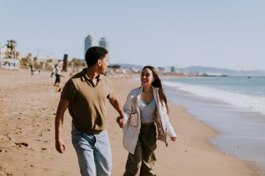 Gülümseyen bir çift kumlu bir sahilde kaygısızca koşmaktan hoşlanır, arka planda yavaşça dalgalar kırılır.