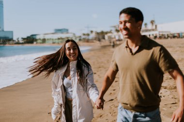 Gülümseyen bir çift kumlu bir sahilde kaygısızca koşmaktan hoşlanır, arka planda yavaşça dalgalar kırılır.