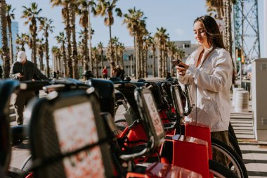 Genç bir kadın güneşli Barcelona 'da bir sıra kırmızı bisikletin yanında duruyor, telefonunu kontrol ediyor, muhtemelen bisiklet paylaşım uygulaması kullanıyor.