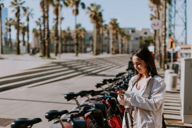 Genç bir kadın güneşli Barcelona 'da bir sıra kırmızı bisikletin yanında duruyor, telefonunu kontrol ediyor, muhtemelen bisiklet paylaşım uygulaması kullanıyor.