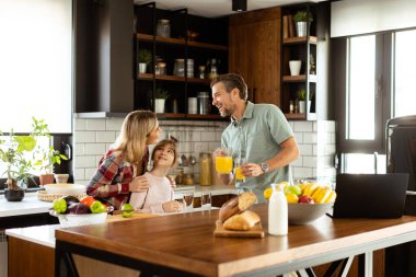 Genç bir aile taze malzemeler ve mutfak malzemeleriyle dolu bir tezgahın etrafında sohbet ediyor ve yemek hazırlıyor.