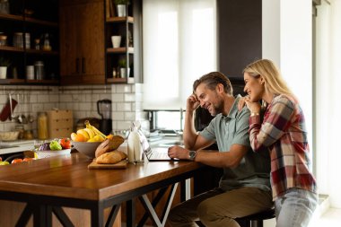 Neşeli bir çift güneşli mutfaklarında keyifli bir anın tadını çıkarıyor. Sağlıklı bir kahvaltıyla çevrili bir laptopla çalışıyorlar.