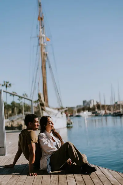Couple sits closely on a dock, enjoying a peaceful moment overlooking the marina on a sunny day in Barcelona.