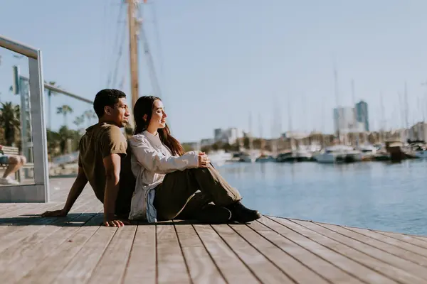 Couple sits closely on a dock, enjoying a peaceful moment overlooking the marina on a sunny day in Barcelona.