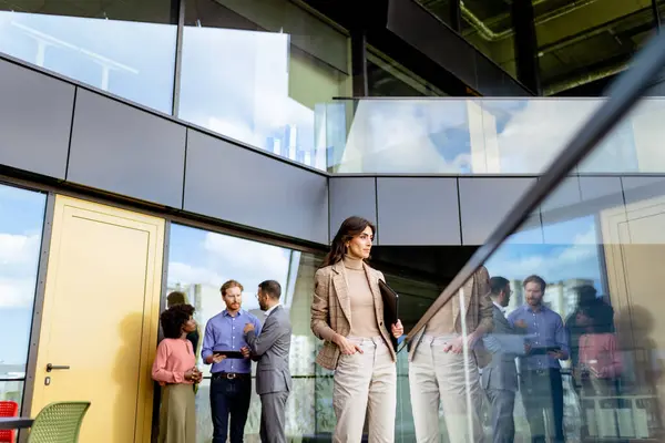 質素なプロの女性がガラス張りのオフィススペースを歩き回り 同僚が背景で会話する ロイヤリティフリーのストック写真