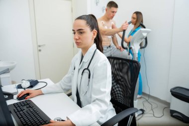 Odaklanmış bir sağlık uzmanı, arka planda hastaları olan bir klinikteki bilgisayardaki verileri inceler.