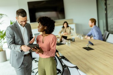 Gülümseyen adam ve Afrikalı Amerikalı iş kadını arka planda iş arkadaşları olan bir tabletin keyfini çıkarıyorlar.